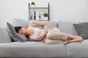 il donna con un' mal di stomaco è Tenere sua stomaco nel dolore, posa su il divano nel il vivente camera. foto