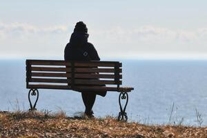 singolo ragazza nel un' nero giacca e cappello seduta su panchina a scogliera a davanti di mare tranquillo, calmo silenzioso posto foto