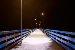 mare molo coperto con neve nel notte giallo luci a partire dal lampioni, misterioso sentiero nel buio foto