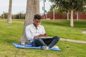 un afroamericano uomo gode il all'aperto seduta nel il erba e utilizzando un' cellula Telefono. foto