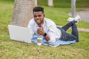 un afroamericano uomo dire bugie nel il verde erba sorridente e pollici su, Lavorando su un' il computer portatile computer. foto