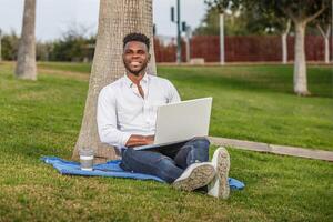 un' sorridente afroamericano uomo seduta sotto un' albero, Lavorando o studiando con un' il computer portatile computer. foto