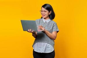 Cheeful giovane donna asiatica che tiene e utilizza il computer portatile isolato su sfondo giallo foto