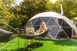 mezzo anziano donna usi un' il computer portatile riposo e la spesa tempo a glamping Casa su vacanze. vacanza cupola tenda. accogliente, campeggio, hygge, stile di vita concetto foto