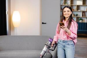 contento allegro giovane caucasico bellissimo donna passare l'aspirapolvere pavimento e sms, fare surf Internet su smartphone a casa nel vivente camera. bella femmina utilizzando vuoto più pulito, domestico concetto foto