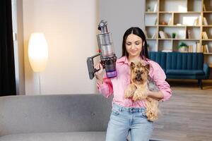 un' carino giovane donna è Tenere un' cordless vuoto addetto alle pulizie con un' divano attaccamento nel uno mano e un' cane nel il altro mano. foto