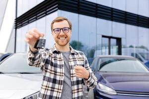 emotivo tipo Tenere chiave nel mano e sorridente a telecamera. uomo mostrando chiave a partire dal il suo nuovo elettrico macchina. giovane uomo acquisto auto a concessionaria salone foto