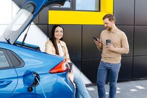 giovane coppia uomo e donna in viaggio insieme di nuovo auto avendo fermare a ricarica stazione. fidanzato Tenere tazza potabile caldo caffè parlando per il Telefono e fidanzata guardare a parte e sorridente foto