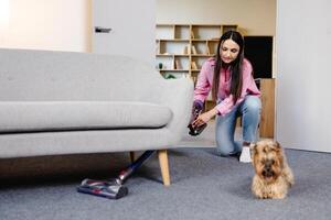 giovane donna usi cordless vuoto addetto alle pulizie per pulito casa tappeto, e un' cane è in esecuzione qui vicino. moderno facile pulizia foto