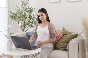 incinta donna Lavorando su il computer portatile e inteligente Telefono nel il vivente camera a casa foto