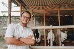 eccitato giovane asiatico uomo in piedi nel davanti di tradizionale gabbia fatto a partire dal legna e bambù nel Indonesia rurale la zona con capra dentro foto