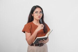 riflessivo giovane asiatico donna indossare un' Marrone camicia e occhiali Tenere un' libro e guardare a parte isolato al di sopra di bianca sfondo. foto