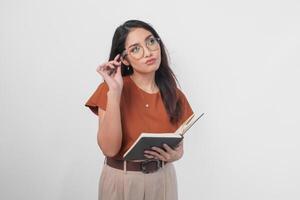 riflessivo giovane asiatico donna indossare un' Marrone camicia e occhiali Tenere un' libro e guardare a parte isolato al di sopra di bianca sfondo. foto