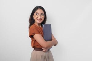 sorridente bellissimo asiatico donna indossare Marrone camicia mentre Tenere documento libro guardare di lato per il vuoto spazio isolato bianca sfondo. foto