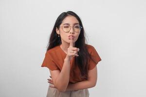 giovane asiatico donna indossare Marrone camicia e monocolo guardare per telecamera mentre mettendo un' dito nel davanti di labbra Esprimere a gesti restare silenzioso o restare silenzioso, isolato di bianca sfondo. foto