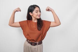 bellissimo asiatico donna indossare un' Marrone camicia in posa forte gesto di sollevamento sua braccia e muscoli mentre sorridente con orgoglio. foto