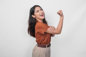 bellissimo asiatico donna indossare un' Marrone camicia in posa forte gesto di sollevamento sua braccia e muscoli mentre sorridente con orgoglio. foto