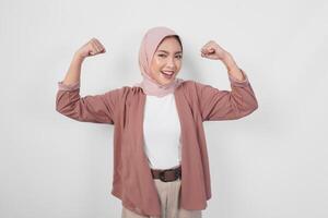 eccitato asiatico musulmano donna indossare un' hijab mostrando forte gesto di sollevamento sua braccia e muscoli sorridente con orgoglio. foto