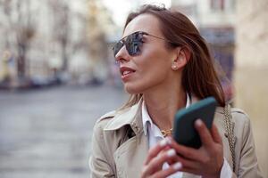 alla moda europeo donna nel occhiali da sole è in piedi con Telefono su città strada sfondo foto