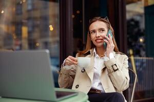 attraente europeo donna parlando Telefono mentre Lavorando in linea seduta a all'aperto bar terrazza foto