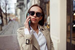 focalizzata donna d'affari nel occhiali da sole è parlando Telefono su città strada sfondo foto