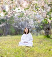 asiatico donna è fare meditazione sotto fiore albero durante il ciliegia fioritura stagione per interno pace, consapevolezza e zen pratica foto