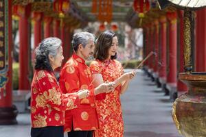 asiatico famiglia con anziano genitore nel rosso cheongsam qipao vestito è offerta incenso per il ancestrale Dio dentro Cinese buddista tempio durante lunare nuovo anno per migliore desiderio benedizione e bene fortuna foto