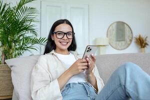 giovane bellissimo donna seduta su il divano e guardare a il telecamera Tenere un' smartphone mobile Telefono, brunetta a casa su il divano nel il vivente camera foto