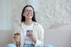 giovane bellissimo donna nel bicchieri sorridente e contento fare in linea shopping nel in linea memorizzare seduta su divano a casa, Tenere mobile smartphone e banca credito carta foto