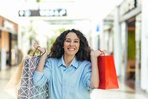 ritratto di un' contento donna acquirente, ispanico donna nel un' supermercato con colorato borse è contento e guardare a il telecamera, dentro un' grande capi di abbigliamento memorizzare foto