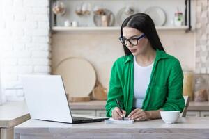 un' giovane grave donna è avendo un' a distanza lavoro colloquio in linea. seduta a casa nel il cucina con un' il computer portatile. parla attraverso un' chiamata, introduce lui stesso, passa il esame, scrive nel un' taccuino. foto