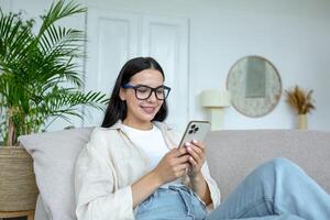 contento e sorridente giovane adolescenziale ragazza dire bugie su divano a casa e utilizzando Telefono. riceve messaggi, chat con gli amici, fidanzato, Chiacchierare notizia, blog, sociale reti, in linea shopping.. foto