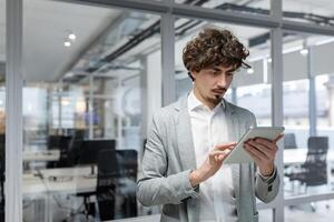 un' poppa pensiero uomo d'affari sta dentro il ufficio con un' tavoletta computer, un' uomo nel un' attività commerciale completo da uomo usi un in linea applicazione. foto