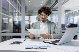 riuscito finanziere investitore lavori con documenti e conti dentro ufficio, uomo d'affari con barba sorridente soddisfatto con opera risultati e risultati, uomo dentro ufficio con il computer portatile Lavorando. foto