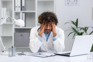 stanco indiano medico Lavorando nel medico ufficio, uomo Tenere mani su testa a partire dal sovraccarico, seduta a scrivania con il computer portatile nel medico bianca cappotto. foto