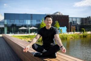 asiatico giovane uomo atleta, allenatore, professionista si siede di il lago su un' panchina nel il loto posizione, medita, fa yoga. sorridente a il telecamera. foto