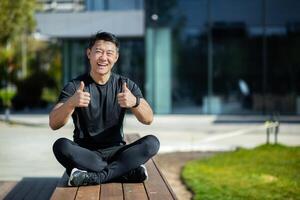 asiatico giovane atleta, allenatore, professionista si siede su il lago riva su un' panchina nel il loto posizione, medita, fa yoga. sorridente a il telecamera, lui Spettacoli il suo dita super, successo. foto