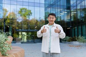 asiatico uomo d'affari, fondatore di un' azienda e azienda in piedi nel davanti di un ufficio centro, sorridente a il telecamera e mostrando super successo segni con il suo dita. foto