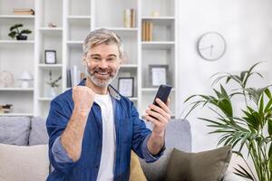 ritratto di un' dai capelli grigi anziano uomo seduta a casa su il divano e utilizzando il Telefono. lui sembra a il telecamera e punti con il suo mano sì, festeggia, gioisce. foto