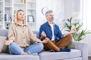 anziano coppia seduta su divano con occhi chiuso nel loto posizione, praticante yoga nel luminosa vivente camera. gli sport a casa, un' salutare stile di vita, calmante e rilassante dopo un' difficile giorno opera. foto