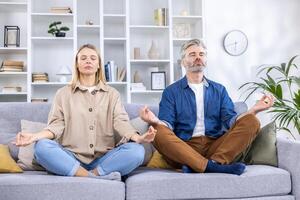 adulto maturo coppia uomo e donna rilassante insieme a casa seduta su divano nel loto posizione, famiglia Meditare insieme nel vivente camera. foto