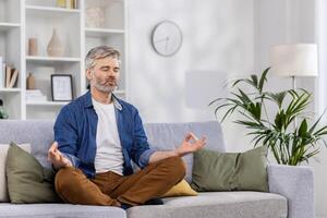 adulto maturo uomo solo a casa rilassante seduta nel loto posizione su divano, grigio dai capelli persona rilassante Meditare in casa nel vivente camera nel casuale Abiti. foto