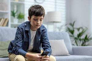 un' bambino di un' adolescenziale ragazzo seduta a casa su il divano e Tenere un' Telefono nel il suo mani. chat, giochi in linea Giochi e applicazioni, usi sociale reti, legge notizia. foto