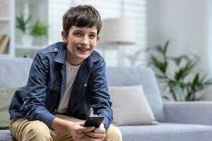 un' bambino di un' adolescenziale ragazzo seduta a casa su il divano e Tenere un' Telefono nel il suo mani. chat, giochi in linea Giochi e applicazioni, usi sociale reti, legge notizia. sorridente guardare a il telecamera. foto