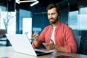 apprendimento in linea. un' giovane maschio insegnante nel un' cuffia e un' rosso camicia si siede nel il ufficio a un' tavolo, lavori su un' computer portatile, insegna attraverso un' chiamata, conferenza, seminario web. foto