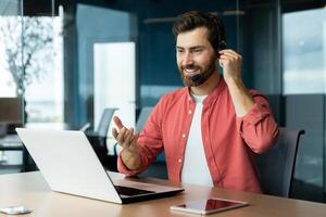 ritratto di un' giovane uomo nel un' cuffia e un' rosso camicia. lui si siede a il scrivania nel il ufficio, lavori su un' computer portatile, comunica attraverso un' chiamata, colloqui, sorrisi. foto