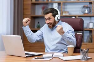 maturo uomo d'affari nel camicia e cuffie Guardando gli sport incontro a posto di lavoro dentro ufficio, uomo applauso avendo divertimento a opera, Guardando su il computer portatile. foto