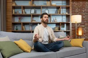 maturo uomo a casa seduta su divano nel sera Meditare nel loto posizione nel vivente camera. foto