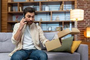 arrabbiato imbrogliato uomo a casa con Telefono e Incorniciato pacco ricevuto, discutere su Telefono parlando per in linea memorizzare cliente supporto servizio, Prodotto consegna fallito. foto