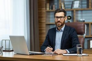 ritratto di riuscito sorridente uomo nel ufficio, maturo uomo d'affari guardare a telecamera allegramente, anziano capo nel bicchieri e barba Lavorando dentro ufficio con il computer portatile e documenti, firma contrarre. foto